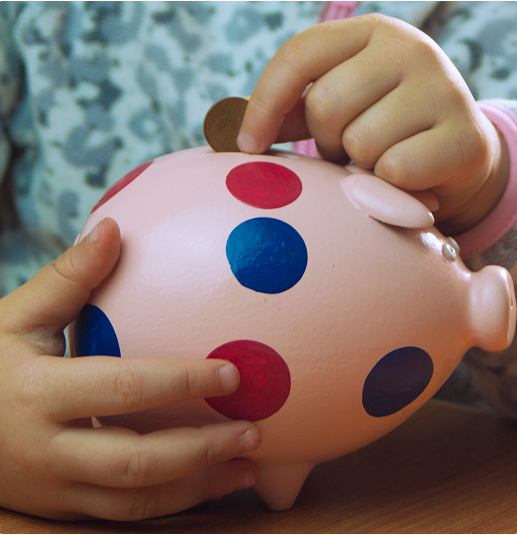 Un enfant insère une pièce de monnaie dans une tirelire rose à pois rouges et bleus, posée sur une table en bois.