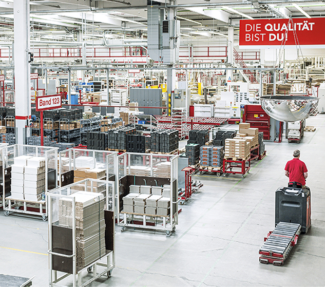 Une usine avec des palettes de cartons empilés, des chariots de transport et un panneau rouge suspendu affichant "DIE QUALITÄT BIST DU!".