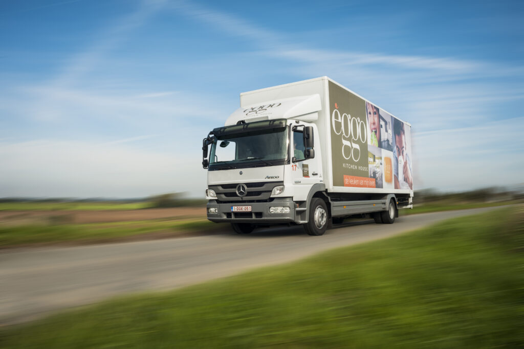 Camion blanc avec logo èggo sur la route, ciel bleu en arrière-plan, champs verts à gauche.