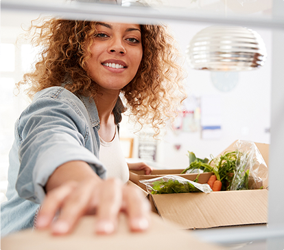 Femme souriante portant une chemise en jean, atteignant une boîte en carton contenant des légumes frais dans une cuisine moderne.