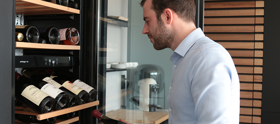 Un homme en chemise bleue regarde des bouteilles de vin dans une cave à vin moderne avec des étagères en bois.