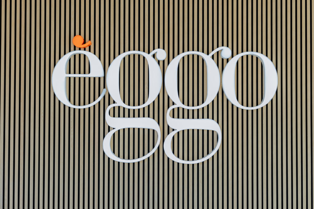 Logo blanc "èggo" sur un fond de lattes en bois verticales, avec un petit oiseau orange posé sur le "e".