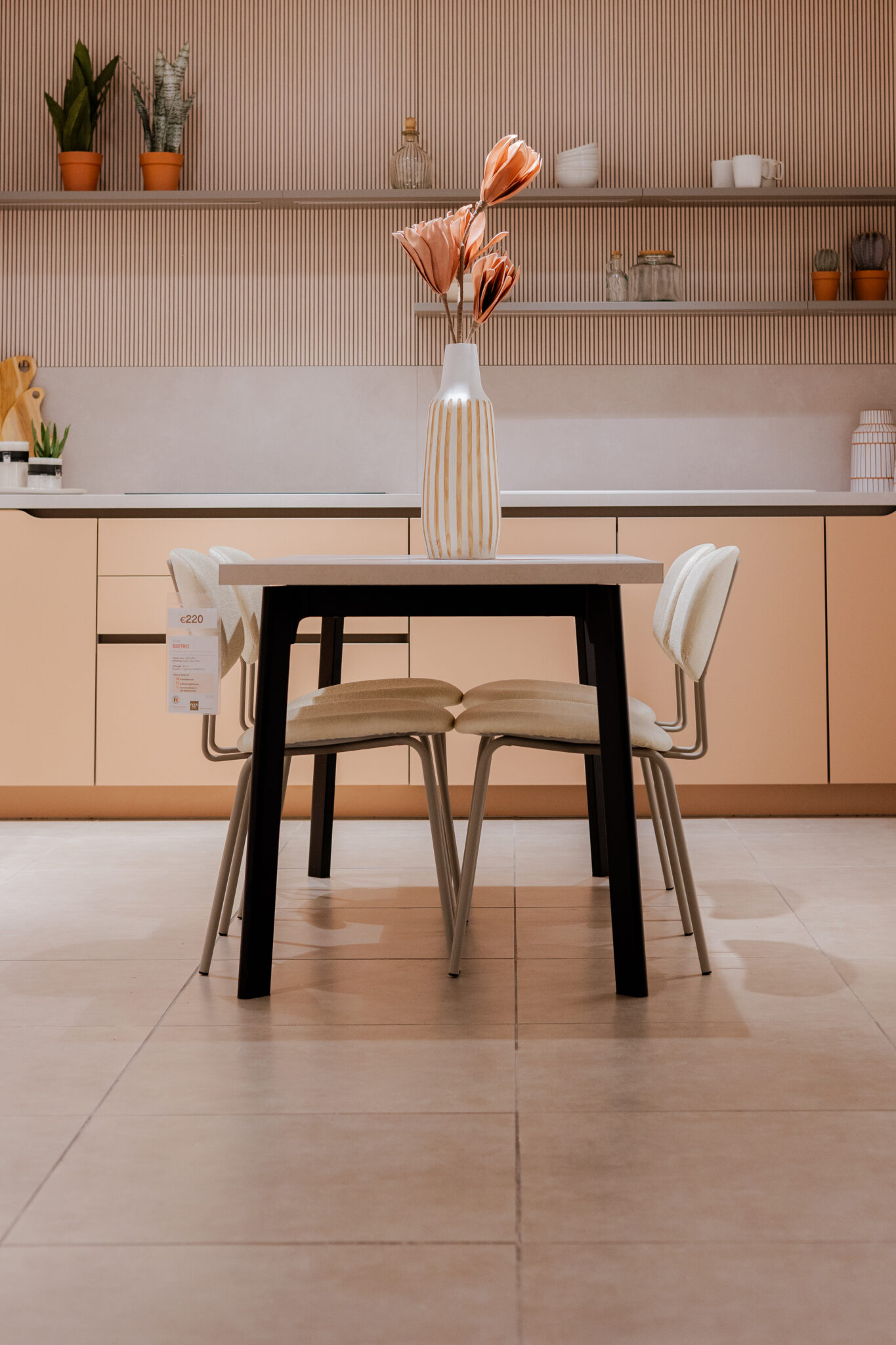 Une table avec un vase à fleurs roses est entourée de chaises beiges dans une cuisine moderne aux tons clairs.