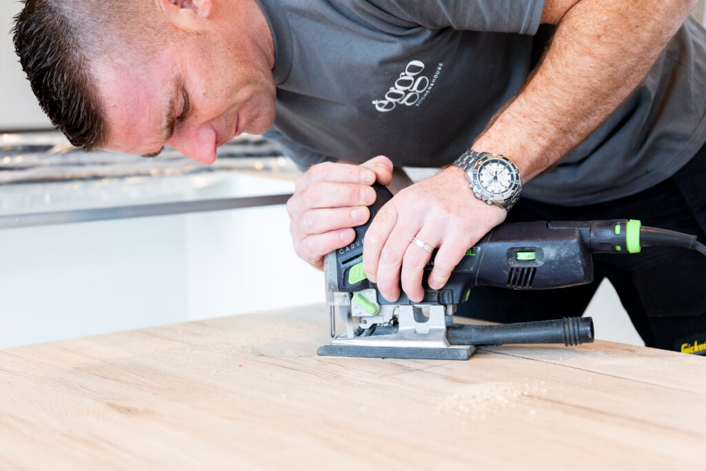 Un homme utilise une scie sauteuse noire et verte sur une planche en bois clair, vêtu d'un t-shirt gris.