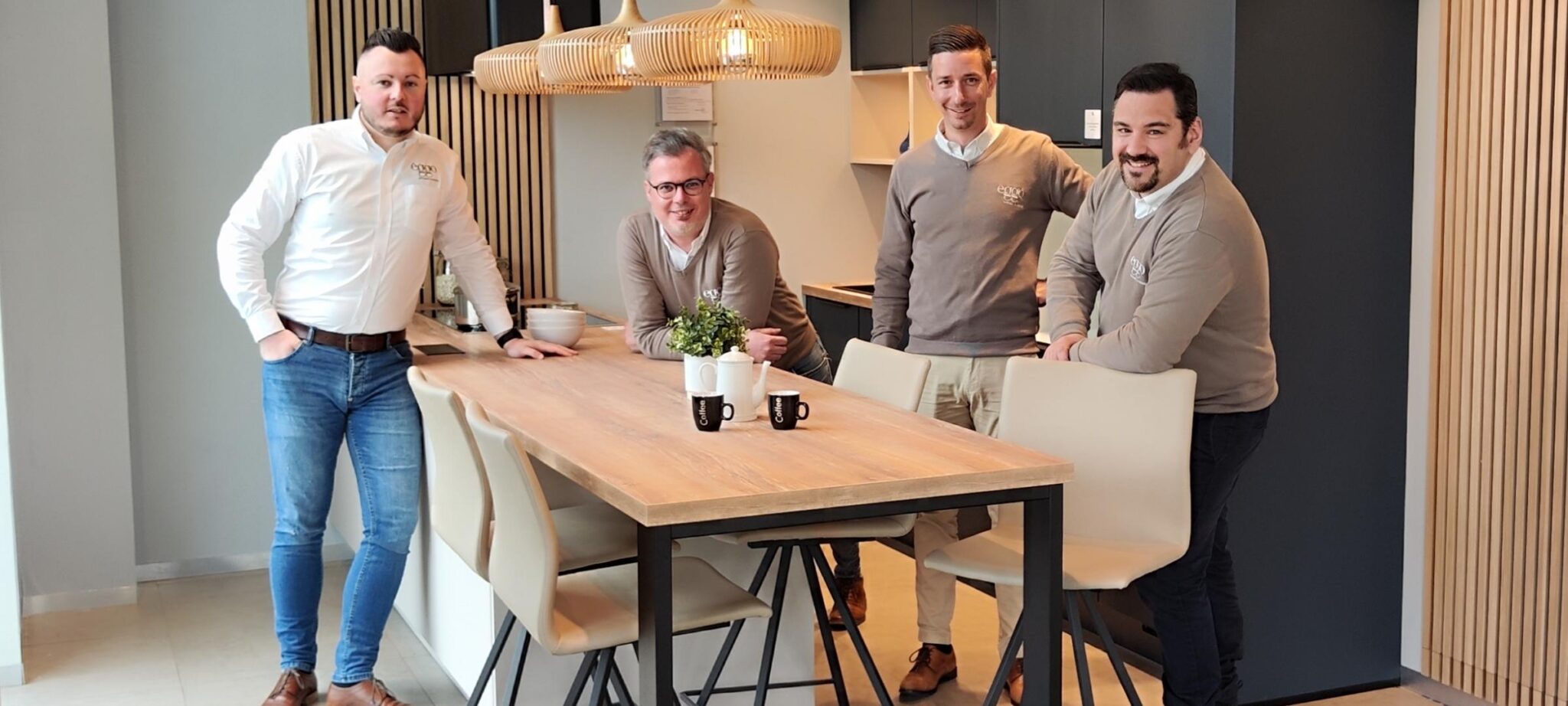 Quatre personnes posent autour d'une table en bois avec des chaises beiges, des tasses noires et une plante verte.