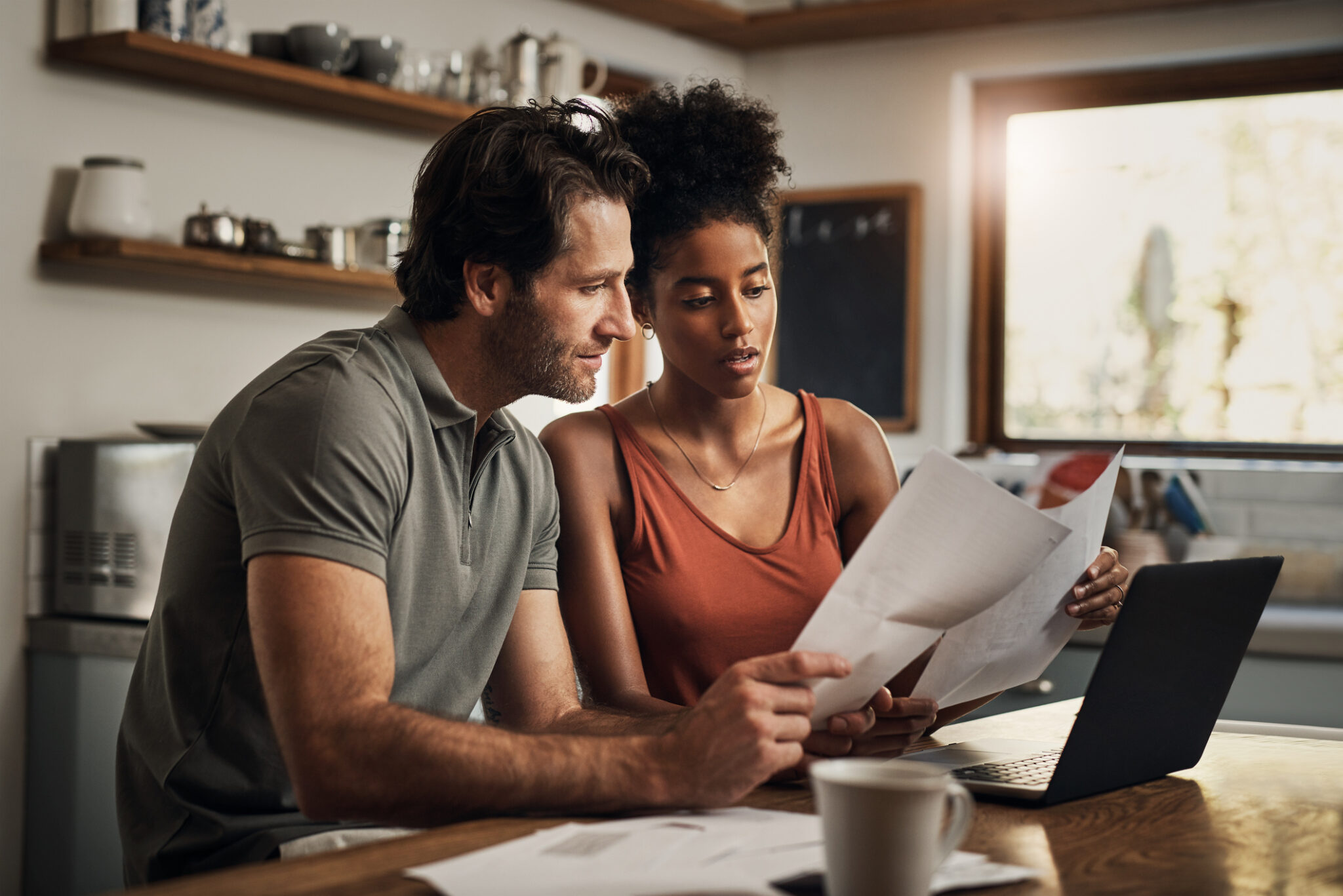 Deux personnes regardent des documents à côté d'un ordinateur portable sur une table en bois dans une cuisine moderne.