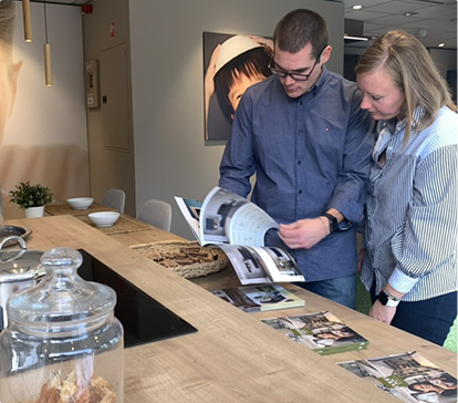 Deux personnes regardent un catalogue sur une table en bois, à côté d'un bocal en verre et des brochures.