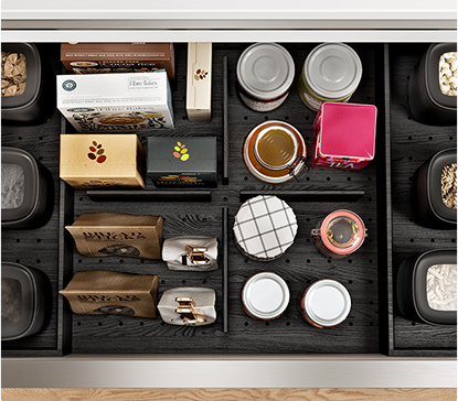 Des bocaux, boîtes et sachets de thé sont organisés dans un tiroir noir compartimenté, avec divers aliments secs et accessoires de cuisine.