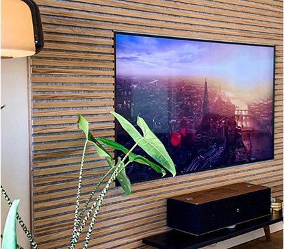 Télévision accrochée sur un mur en bois à lattes, affichant une ville au coucher du soleil, avec des plantes vertes au premier plan.