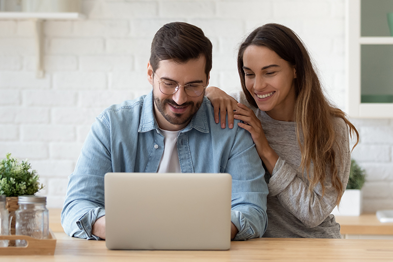 Un homme portant une chemise en jean et une femme en pull gris sourient en regardant un ordinateur portable sur une table en bois.
