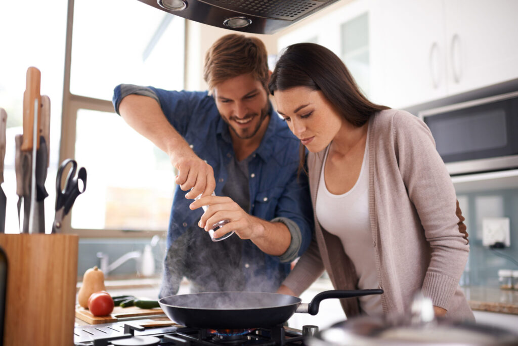 Un homme et une femme sont penchés au-dessus d'une poêle, en train de cuisine. L'homme utilise un moulin à poivre.