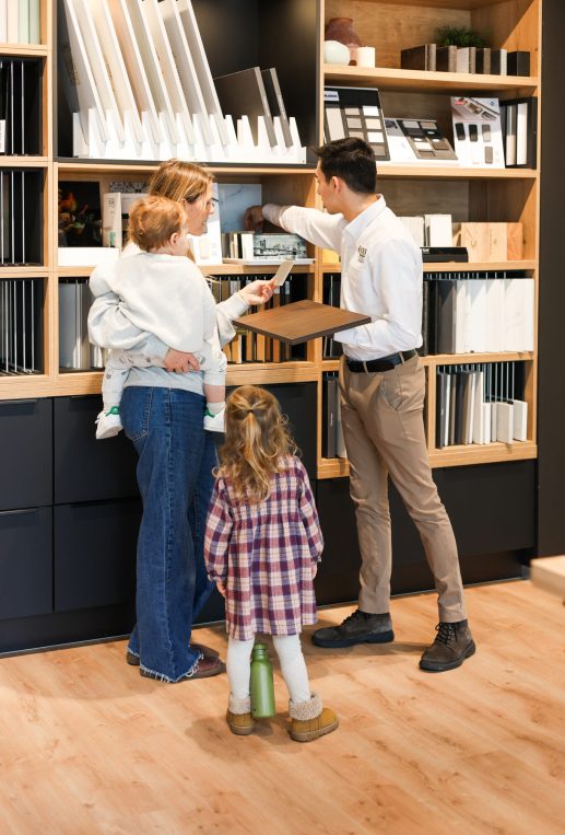 Une femme avec deux enfants consulte un conseiller dans un magasin de cuisines, entourée d'échantillons de matériaux et de couleurs.